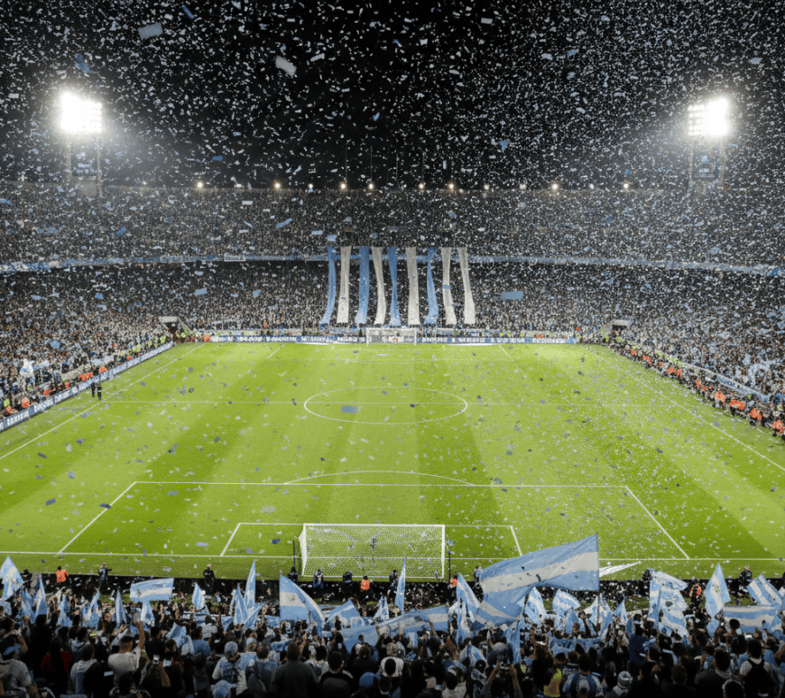 Estadio de fútbol lleno de noche con banderas celestes y blancas y papel picado