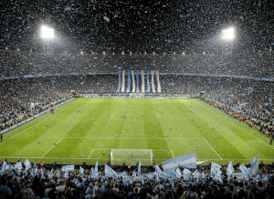 Estadio de fútbol lleno de noche con banderas celestes y blancas y papel picado