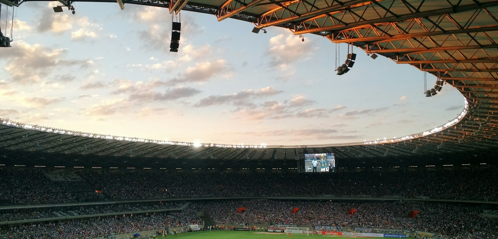 Hinchas en las tribunas de un estadio de fútbol bajo el techo, con la cancha al fondo
