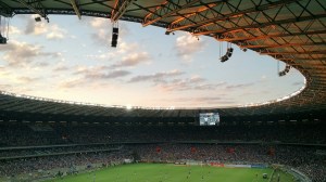 Hinchas en las tribunas de un estadio de fútbol bajo el techo, con la cancha al fondo