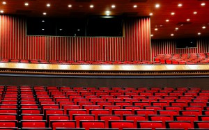 Auditorio vacío con butacas rojas, palcos y un escenario iluminado